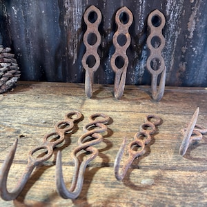 May include: Several rusty metal hooks with a decorative chain-link design are arranged on a wooden surface. The hooks are various sizes and shapes, with a weathered appearance. A pinecone is visible in the background.