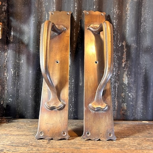 Puede incluir: Dos manijas de puerta antiguas de latón. Cada manija está montada sobre una placa rectangular con bordes decorativos. Las manijas tienen un diseño curvo y una pátina cálida y envejecida.