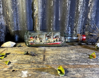Op de afbeelding: Een miniatuur scheepsmodel in een heldere glazen fles met een kurkstop. De fles ligt op een houten oppervlak met een dennenappel en andere natuurlijke elementen.