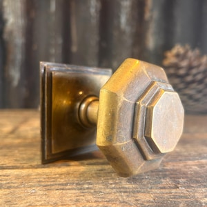 Pode incluir: Um puxador de porta de latão vintage com uma placa traseira quadrada e um puxador octogonal. O puxador tem uma aparência desgastada, sugerindo idade e uso. O fundo é uma superfície de madeira rústica.