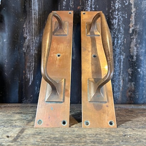 May include: Two rectangular brass door plates with attached arched handles. Each plate features a weathered, metallic finish with visible screw holes at the top and bottom for mounting. The handles are curved and centered on the plates.