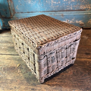 Peut inclure: Une boîte rectangulaire en osier tressé avec un couvercle assorti. La boîte présente une finition brun clair patinée, avec un motif de tressage serré et complexe sur le dessus et un design vertical plus ouvert sur les quatre côtés.