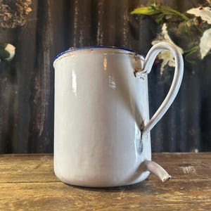 May include: White enamelware jug with a curved handle and a spout near the base. The rim is trimmed with blue. The jug has a vintage, rustic appearance, suggesting it is an antique or a reproduction of an antique.