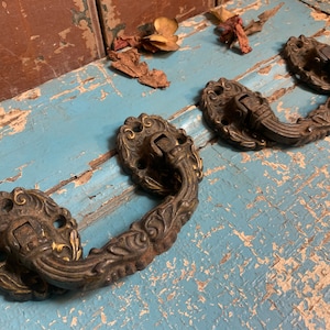 Op de afbeelding: Drie sierlijke metalen ladegrepen met een decoratief bloemmotief. De grepen zijn donkerbrons van kleur en hebben een licht verweerde afwerking.