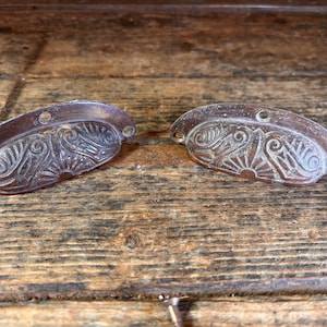 May include: Two antique bronze-colored drawer pulls with ornate, symmetrical designs. Each pull has a curved, half-moon shape with a decorative pattern of swirls and sunburst-like elements. The pulls have two holes for mounting.