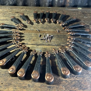 Puede incluir: Una colección de veinticuatro tiradores de cajón de metal dispuestos en círculo, cada uno con un acabado negro oscuro con detalles en tono cobre en los extremos y un diseño de flor de lis. Un pequeño montón de tornillos de montaje a juego se encuentra en el centro.