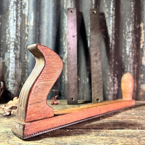 May include: A wooden hand plane with a red-stained finish and a metal blade. The plane has a curved handle and a serrated edge. The plane is sitting on a wooden surface.