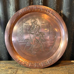 Puede incluir: Una bandeja de metal color cobre con borde elevado. La bandeja presenta una imagen en relieve de un hombre con sombrero de copa y abrigo, caminando con un bast&oacute;n. El texto incluye "Born Eighteen Twenty" y "Still Going Strong."