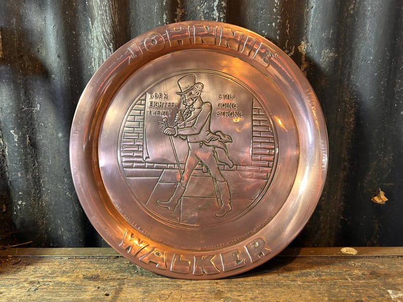 Puede incluir: Una bandeja de metal color cobre con borde elevado. La bandeja presenta una imagen en relieve de un hombre con sombrero de copa y abrigo, caminando con un bast&oacute;n. El texto incluye "Born Eighteen Twenty" y "Still Going Strong."