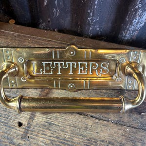 May include: A rectangular brass mail slot plate featuring a central horizontal opening with the word LETTERS embossed in capital letters. The plate includes a large, curved brass handle attached to the front, with decorative circular accents.