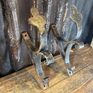 May include: Two antique black metal hooks with ornate gold-colored tops. The hooks have a curved design and are mounted on a wooden surface. Each hook has two holes for mounting. The hooks are likely for hanging items.