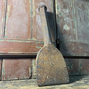 May include: A rusty, metal, hand-held tool with a long, rectangular blade and a rounded handle. The tool is resting on a wooden surface.