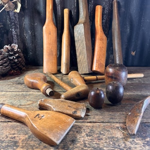 May include: A collection of antique wooden tools, including mallets, paddles, and bottle-shaped implements, arranged on a weathered wooden surface. The tools vary in size and shape, with a warm, natural wood tone. Some tools have a dark stain, and one paddle has the letter "M" on it.