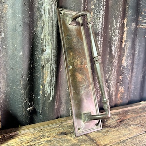 Puede incluir: Un tirador de puerta de latón vintage con una placa trasera rectangular y una larga manija curva. La manija tiene un diseño decorativo y la placa trasera tiene una pátina desgastada y envejecida.