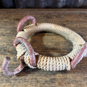 May include: A rustic rope and metal ring with three red-painted metal hooks. The rope is beige and tightly wound, forming a circular shape. The metal shows signs of wear, adding to its vintage aesthetic.