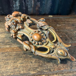 Könnte beinhalten: Ein metallischer Schubladengriff in Bronzetönen mit einem komplexen Reliefdesign aus skulptierten Blättern und floralen Elementen. Das Beschlagteil hat ein zentrales ovales Medaillon mit Perlenrand, ein spitzes Ende und eine verwitterte Patina.