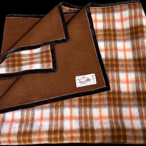 May include: A folded blanket with a brown exterior and a plaid interior in shades of white, orange, and brown. The blanket has a dark brown border and a label in the corner.