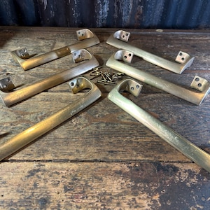 May include: A set of four matching brass drawer pulls with a smooth, elongated design and curved ends. Each handle features two mounting points with pre-drilled holes for screws. A small pile of metal screws is positioned in the center of the group.