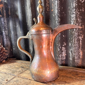 May include: A copper-colored, antique coffee pot with a long spout and ornate handle. The pot has a decorative finial on the lid and sits on a wooden surface. The background features a corrugated metal sheet.