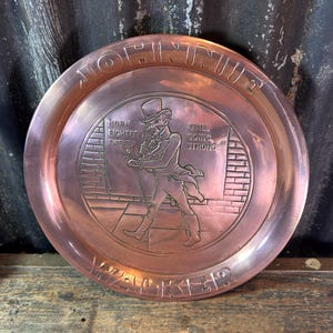 Puede incluir: Una bandeja redonda de metal color cobre con las palabras "JOHNNIE WALKER" alrededor del borde. El centro presenta una imagen en relieve de un hombre con sombrero de copa y abrigo, con el texto "BORN EIGHTEEN TWENTY" y "STILL GOING STRONG".