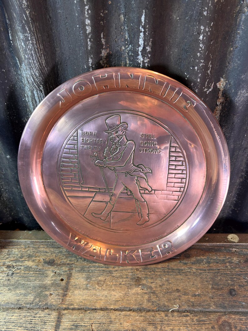 Puede incluir: Una bandeja redonda de metal color cobre con las palabras "JOHNNIE WALKER" alrededor del borde. El centro presenta una imagen en relieve de un hombre con sombrero de copa y abrigo, con el texto "BORN EIGHTEEN TWENTY" y "STILL GOING STRONG".
