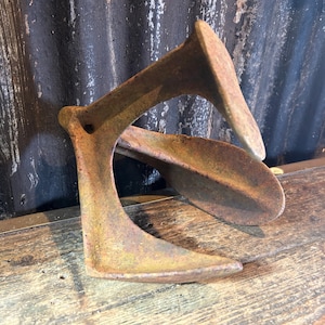 May include: A weathered metal shoe last, showing signs of age and rust. The shoe last has a light brown hue and a distinctive curved form. It rests on a wooden surface, likely a workbench.