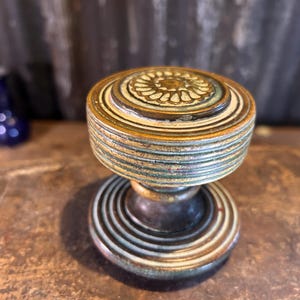 May include: A vintage-style cabinet knob with a round, layered design. The knob features a central floral pattern in cream and is surrounded by concentric rings in shades of gold, green, and bronze. The knob is likely made of metal and has an aged appearance.