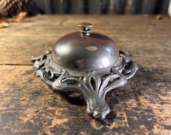 May include: A silver bell with a decorative cast iron base. The bell has a small knob on top and a smooth, rounded surface.
