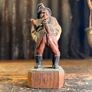 以下が含まれることがあります： 帽子をかぶってパイプを持っている男性の木製人形。人形は茶色、赤、黒の色合いで塗られています。木製の台座の上に立っています。