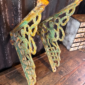 Op de afbeelding: Twee antieke gietijzeren plankdragers met een verweerde groen en gele afwerking. Elke beugel heeft een decoratief ontwerp met een figuur en krullen. De beugels zijn ontworpen om aan de muur te worden bevestigd en een plank te ondersteunen.