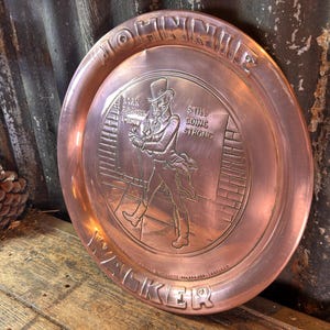 Puede incluir: Una bandeja de metal color cobre con un dise&ntilde;o en relieve. La bandeja presenta una imagen central de un hombre con sombrero de copa y abrigo, con el texto "MORNING WALKER" y "STILL GOING STRONG". La bandeja es redonda y tiene un borde elevado.