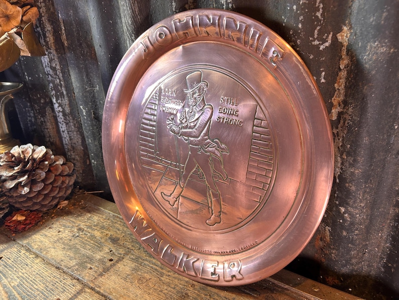 Puede incluir: Una bandeja de metal color cobre con un dise&ntilde;o en relieve. La bandeja presenta una imagen central de un hombre con sombrero de copa y abrigo, con el texto "MORNING WALKER" y "STILL GOING STRONG". La bandeja es redonda y tiene un borde elevado.