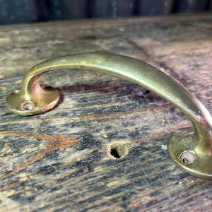 May include: A brass door handle with a curved design. The handle is mounted on a wooden surface.