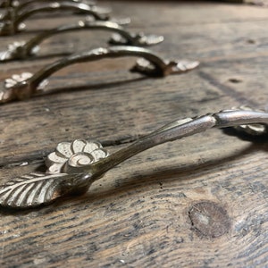 Könnte beinhalten: Sechs silberne Schubladengriffe mit floralem Design. Die Griffe haben eine gebogene Form und sind aus Metall. Die Griffe sind in einer Reihe auf einer Holzoberfläche angeordnet.
