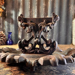 Puede incluir: Soporte decorativo de metal marrón oscuro y ornamentado con un diseño detallado y enrollado. El soporte tiene un soporte central y una base circular con un patrón en forma de hoja. El fondo incluye una pared de metal corrugado y otros elementos decorativos.
