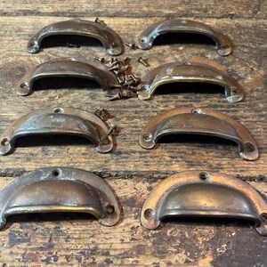 May include: A set of eight semi-circular brass drawer pulls with a weathered, tarnished finish. Each pull features two mounting holes at the ends and a curved handle grip. A small pile of matching metal screws is included with the hardware.