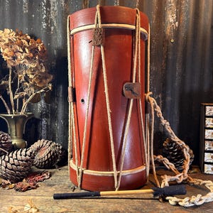 May include: A red wooden drum with a rope lacing system and a wooden stick. The drum is sitting on a wooden surface with pine cones and dried flowers.