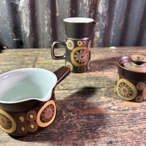 May include: A set of three brown ceramic kitchenware pieces with a floral pattern. The pieces include a creamer, a sugar bowl with a lid, and a small jug.