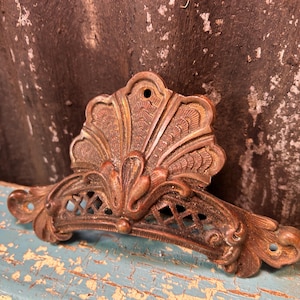 May include: Ornate, antique bronze-colored metal drawer pull with a fan-shaped top and decorative filigree. The pull has a curved handle with diamond-shaped cutouts and is likely from a vintage cabinet or furniture piece.