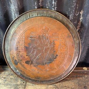 Puede incluir: Una bandeja redonda de metal color cobre con un borde elevado. La bandeja presenta una imagen en relieve de un hombre con sombrero de copa y traje, caminando con un bast&oacute;n frente a una pared de ladrillos. El texto est&aacute; en relieve alrededor del borde y dentro de la imagen.