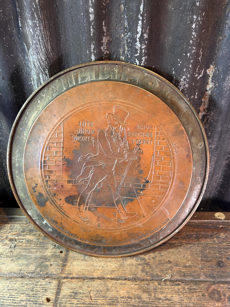 Puede incluir: Una bandeja redonda de metal color cobre con un borde elevado. La bandeja presenta una imagen en relieve de un hombre con sombrero de copa y traje, caminando con un bast&oacute;n frente a una pared de ladrillos. El texto est&aacute; en relieve alrededor del borde y dentro de la imagen.