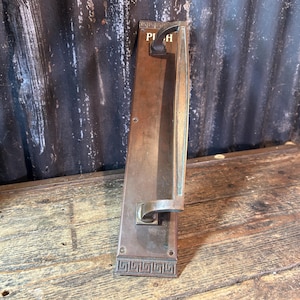 Puede incluir: Una placa de empuje para puerta rectangular de bronce con un asa vertical y la palabra PUSH grabada cerca de la parte superior. La placa tiene una pátina oscura y desgastada y un patrón de greca decorativo en el borde inferior con tornillos.