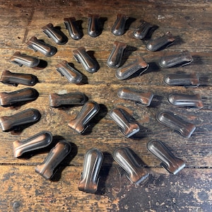 May include: An overhead shot displays a collection of antique bronze-colored metal clips arranged on a weathered wooden surface. The clips have a curved, handle-like design. Additional metal pieces are scattered nearby.