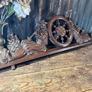 May include: A carved wooden decorative piece featuring two figures playing instruments, a wheel, and ornate details. The figures are seated and dressed in traditional attire. The piece is a warm brown color and rests on a wooden base.