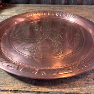Puede incluir: Un plato de metal color cobre con un dise&ntilde;o en relieve. La imagen central representa una figura con abrigo largo y botas, caminando. El borde del plato tiene texto y un borde decorativo. El plato es redondo y tiene un acabado pulido.