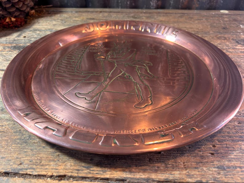 Puede incluir: Un plato de metal color cobre con un dise&ntilde;o en relieve. La imagen central representa una figura con abrigo largo y botas, caminando. El borde del plato tiene texto y un borde decorativo. El plato es redondo y tiene un acabado pulido.