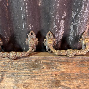 Op de afbeelding: Twee antieke messing ladehandgrepen met sierlijke bloemen- en krulwerkontwerpen. Elke handgreep heeft een sleutelgat en een gebogen handvat. De handgrepen worden getoond op een verweerd houten oppervlak tegen een donkere, getextureerde achtergrond.