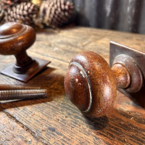 May include: Two antique bronze door knobs with square backplates. The knobs are a dark brown colour with a worn finish. The knobs are on a wooden surface.