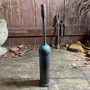 May include: A vintage blue and rusty metal grease gun with a long metal nozzle. The grease gun is sitting on a wooden surface with fallen leaves.