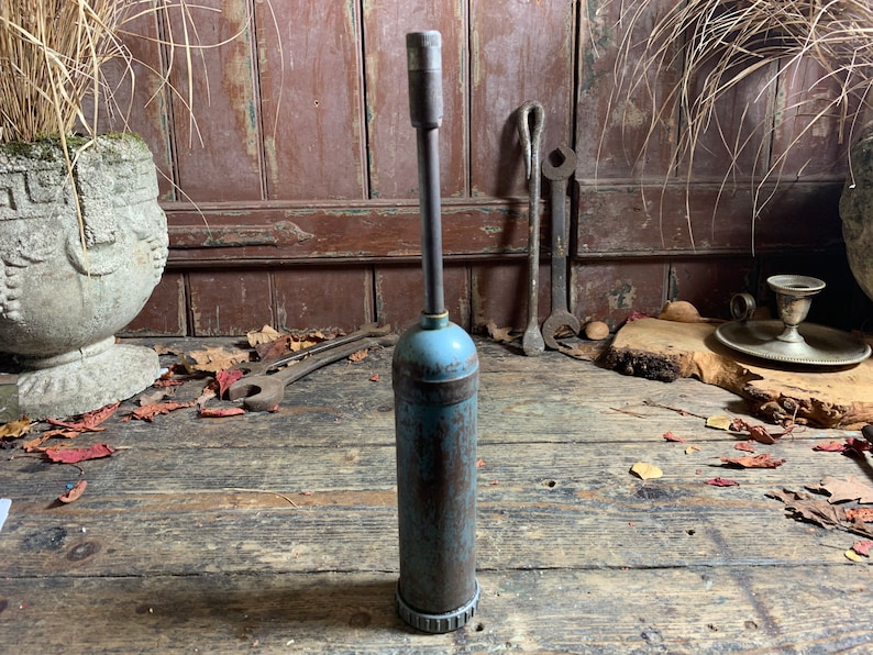 May include: A vintage blue and rusty metal grease gun with a long metal nozzle. The grease gun is sitting on a wooden surface with fallen leaves.